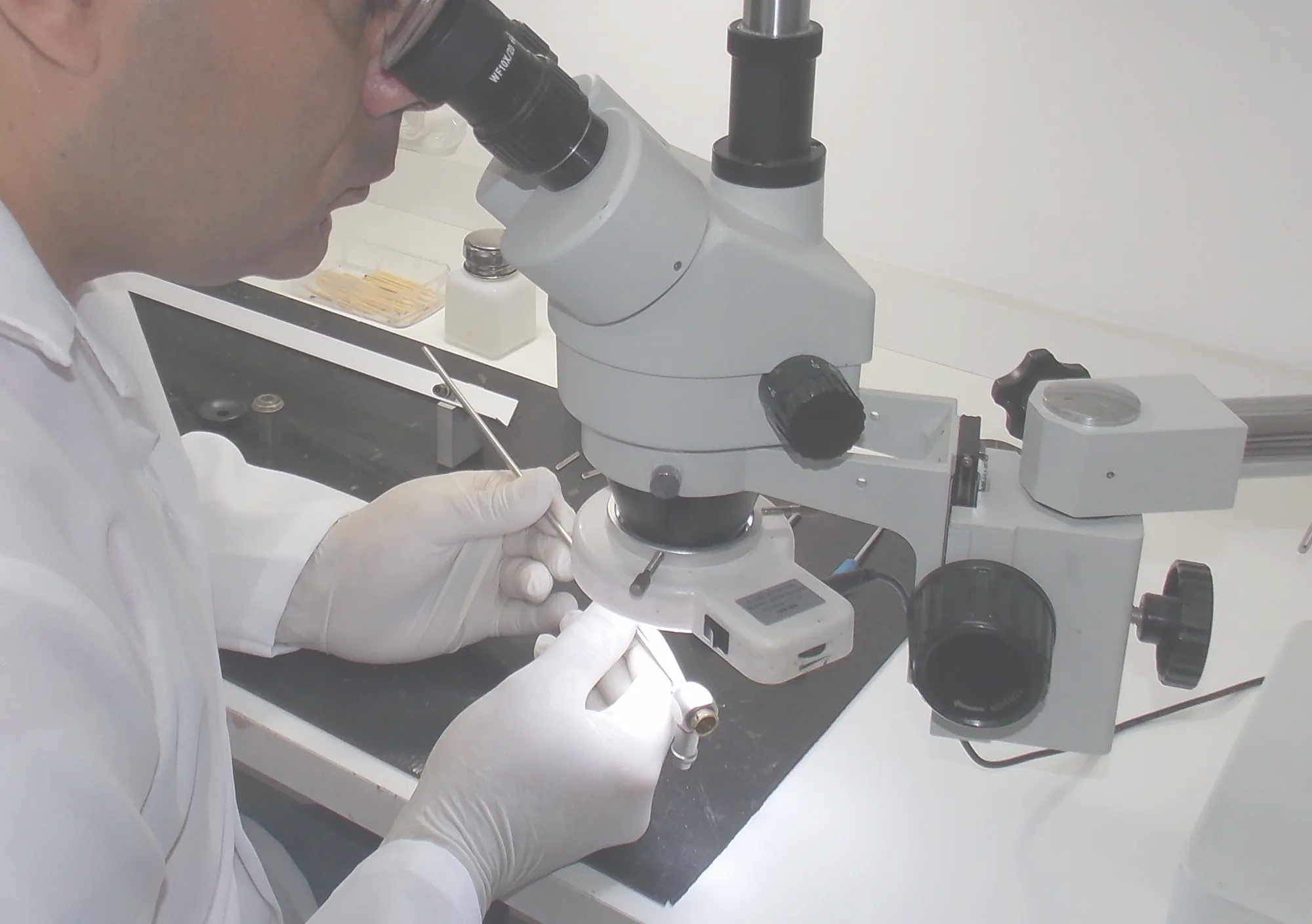 Técnico da AJP Master, de jaleco e luvas, repara uma peça de vídeo cirurgia sob microscópio estereoscópico na bancada da oficina.